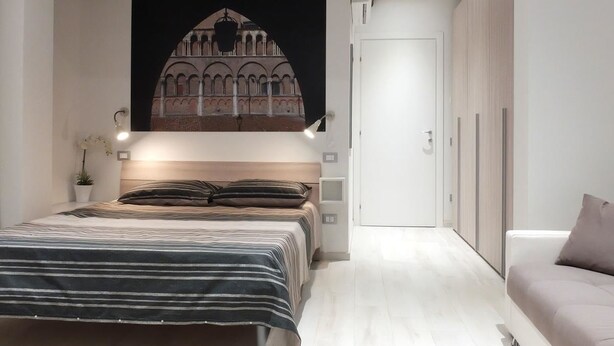 A bedroom with a bed, a nightstand, a wall-mounted lamp, a wardrobe, and a painting of a building. - Centro Storico Suites (Ferrara)