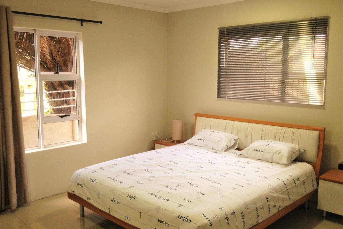 A bedroom with a bed, a nightstand, a window with blinds, and a curtain.