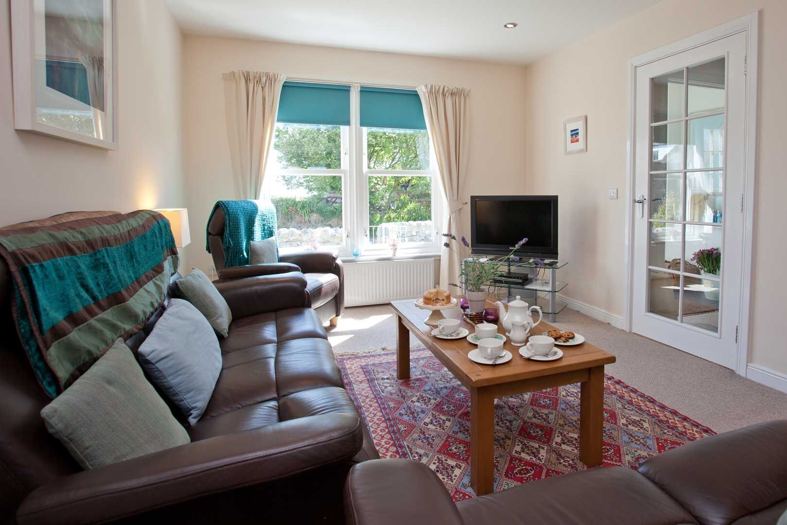 A living room with a brown leather sofa, a wooden coffee table with tea cups and pastries, a television, and a view of greenery outside.