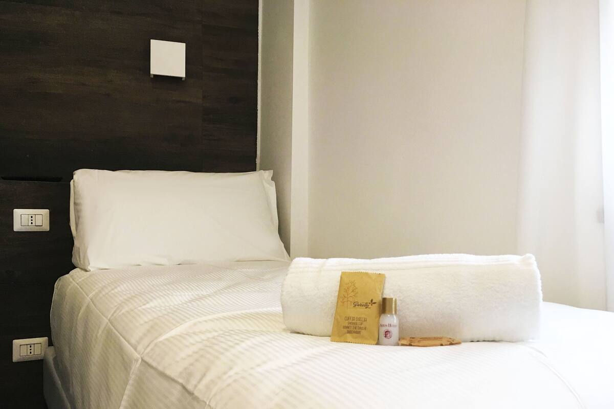 A neatly made bed with a white comforter, a white pillow, and a folded towel. There is a small brown bag and a bottle of lotion on the bed.