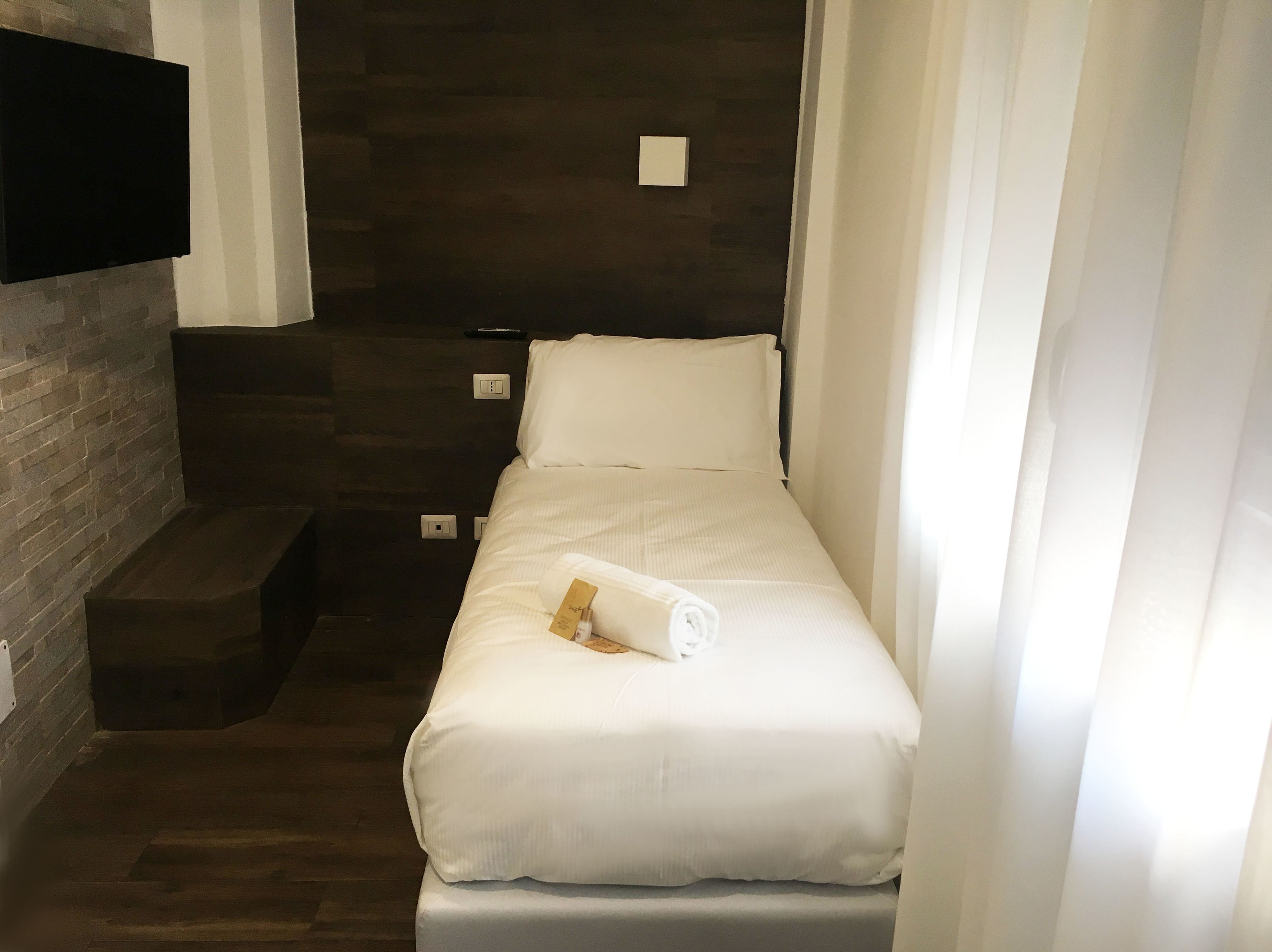 A small, neatly made bed with white bedding and a rolled towel, positioned in a corner with a dark wooden wall and a small step leading up to it.