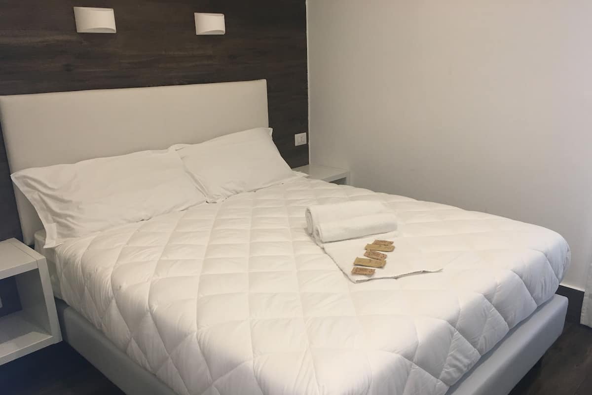 A neatly made bed with a white quilted duvet, a rolled white towel, and a few brown slippers on the bed.