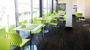 A modern restaurant with green chairs and white tables, large windows, and a dark wooden floor.