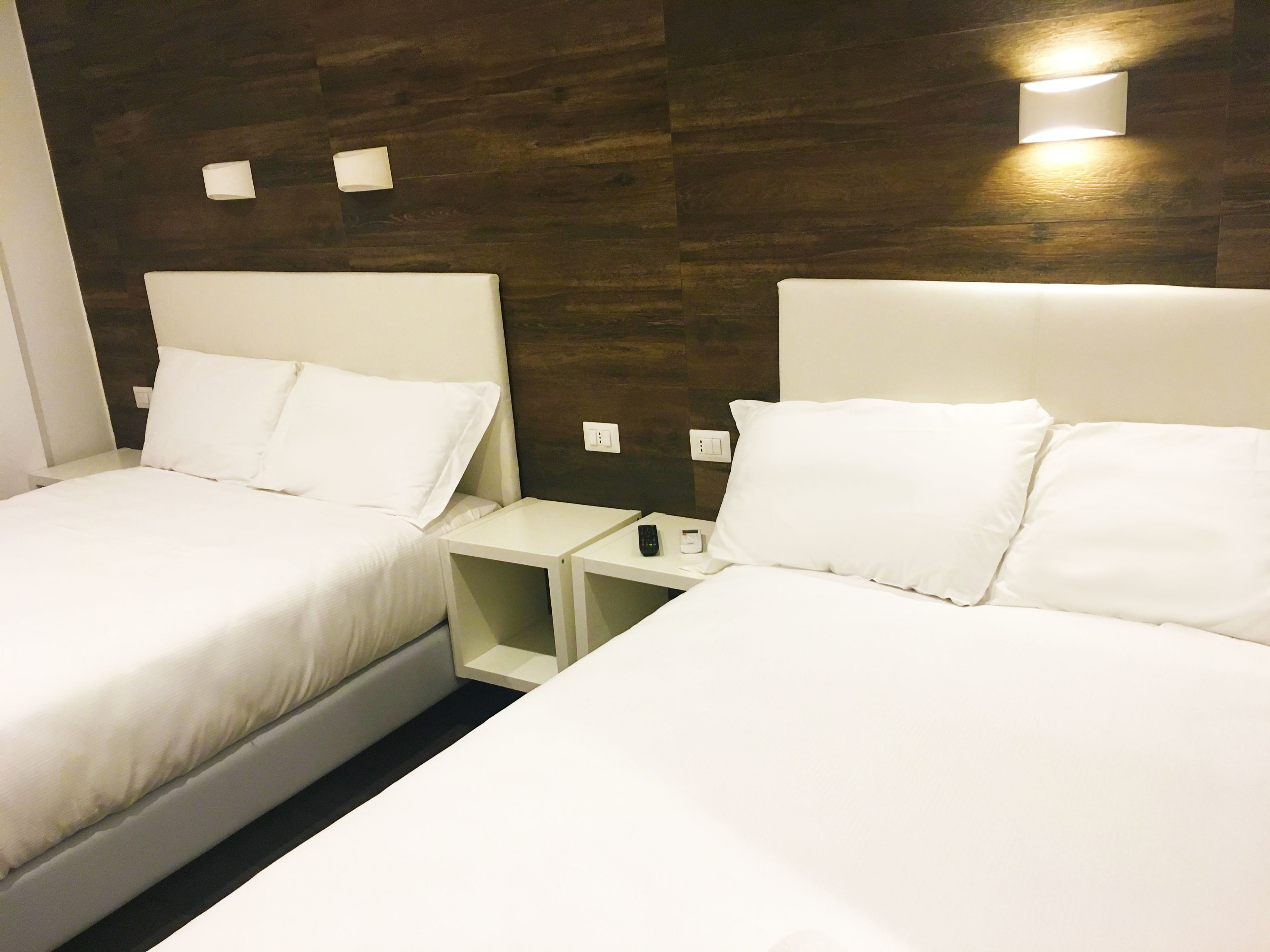 Habitación de hotel con dos camas, cada una con sábanas y almohadas blancas. Mesitas de noche con lámparas y un control remoto. Paredes de madera con acabado oscuro.