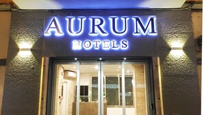 The exterior of AURUM HOTELS with a prominent illuminated sign, large glass doors, and a stone facade.