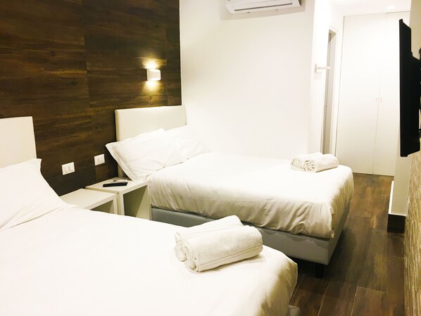 Habitación de hotel con dos camas, una mesita de noche, aire acondicionado en la pared y una puerta que da a otra habitación.