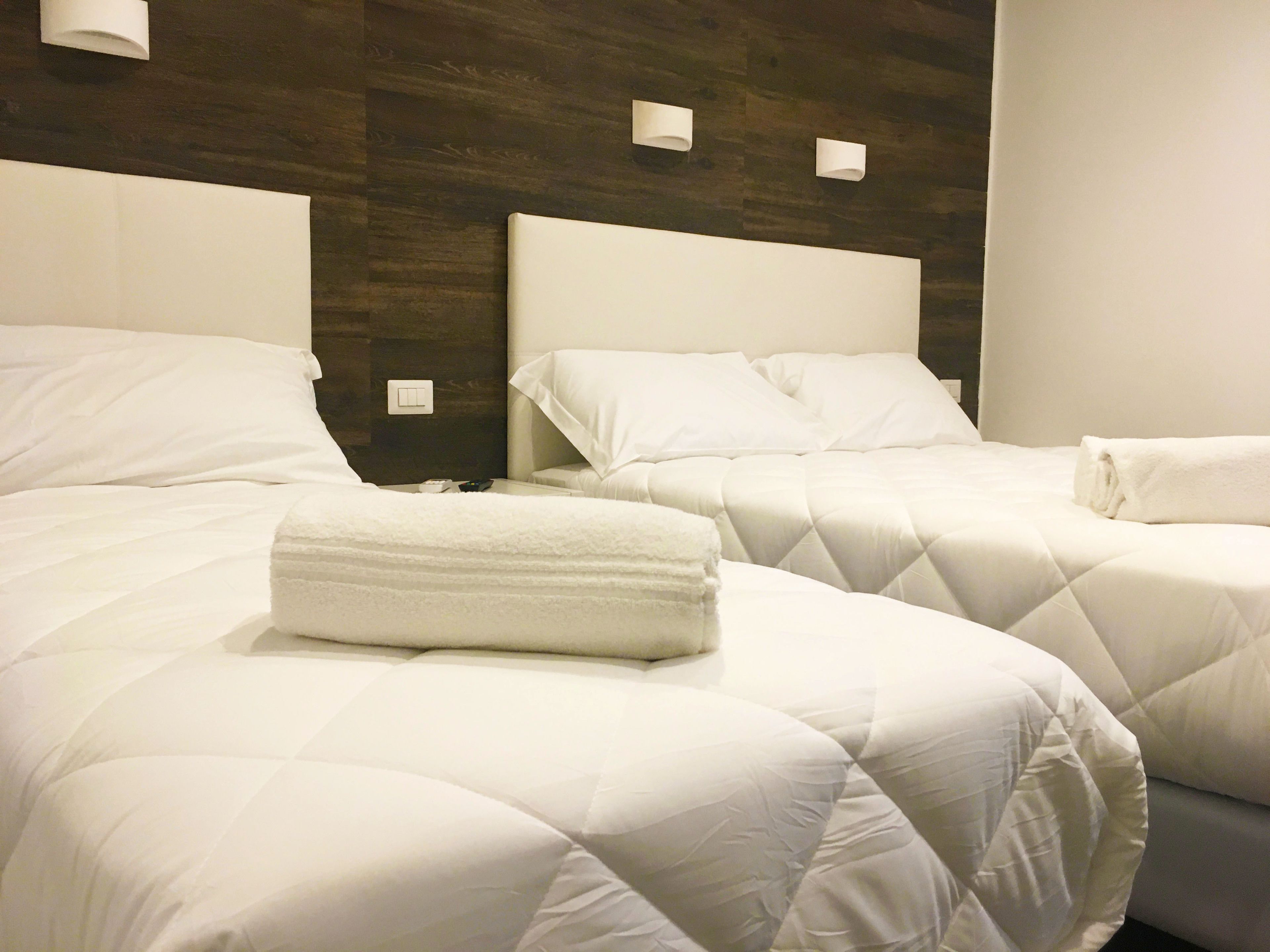 A hotel room with two beds, each with white bedding and a quilted duvet. There are two bedside tables with lamps and a wall-mounted light fixture.