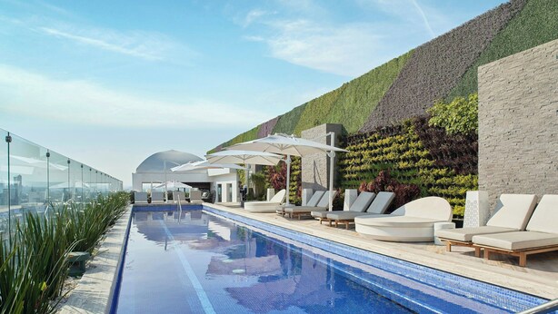 Een dakterraszwembad met ligbedden en parasols, omgeven door een aangelegde tuin en een modern gebouw.