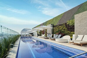 Een dakterraszwembad met ligbedden en parasols, omgeven door een aangelegde tuin en een modern gebouw.