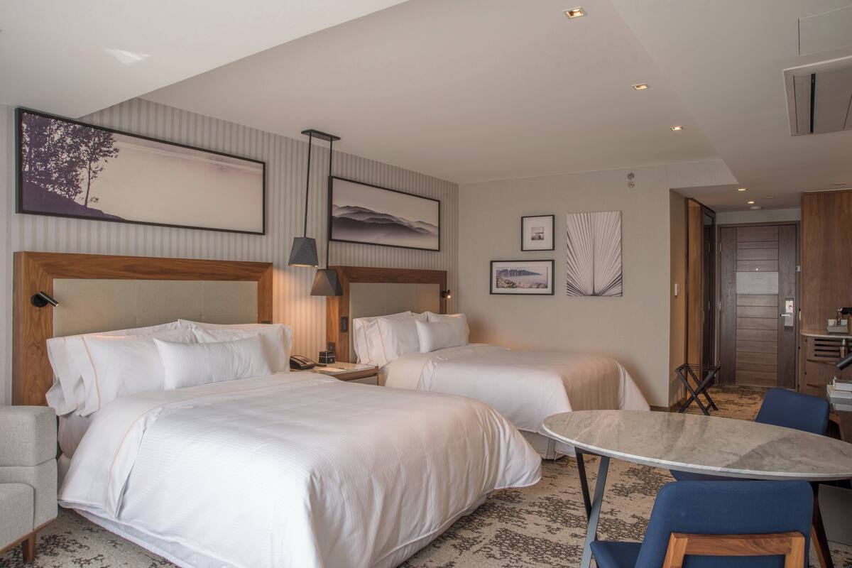 Une chambre d’hôtel avec deux lits, une petite table et une chaise. Les murs sont ornés de tableaux encadrés et une lampe se trouve sur la table de chevet.