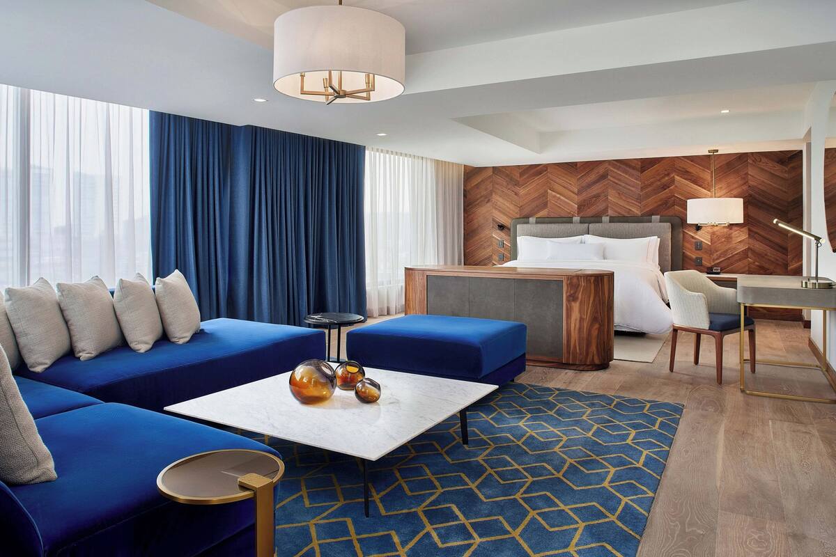Une chambre d’hôtel moderne avec un canapé bleu, une table basse blanche, un mur en bois à motifs en chevrons, un lit avec une tête de lit grise et un bureau avec une lampe.