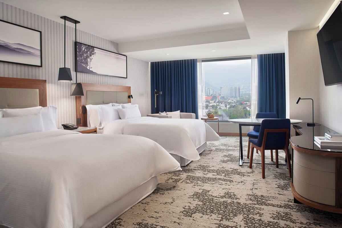 Une chambre d’hôtel avec deux lits, un bureau, une chaise, une télévision et une grande fenêtre donnant sur la ville.