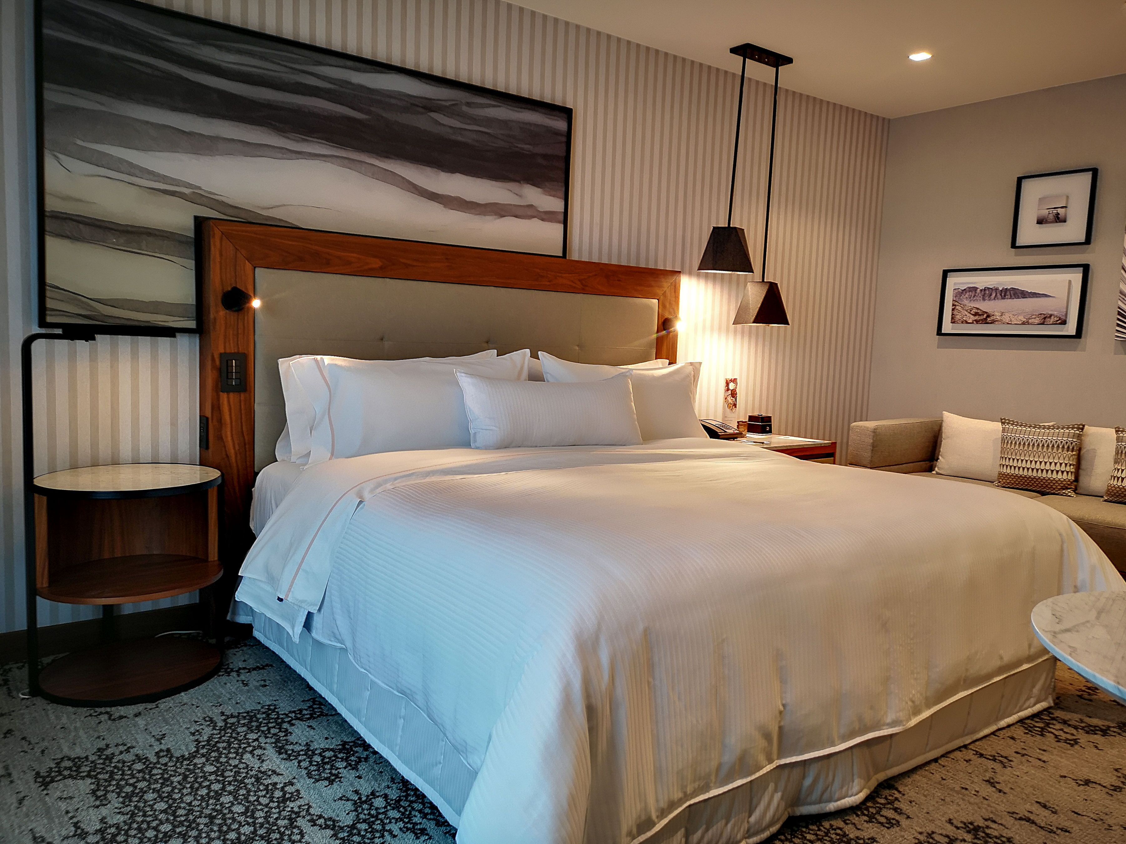 Une chambre d’hôtel avec un grand lit, une petite table ronde, un canapé, un téléviseur fixé au mur et un tableau.
