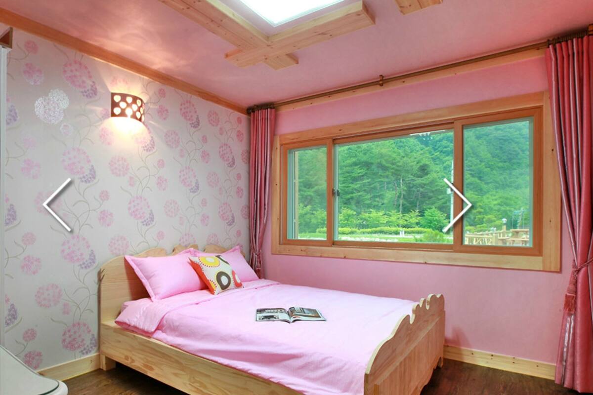 A bedroom with a wooden bed, pink bedding, a window with a view of greenery, and a wall-mounted light fixture.