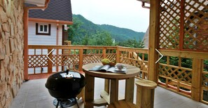 A wooden deck with a barbecue grill, a table with a bowl of fruit and a teacup, and a view of a house and mountains in the background.
