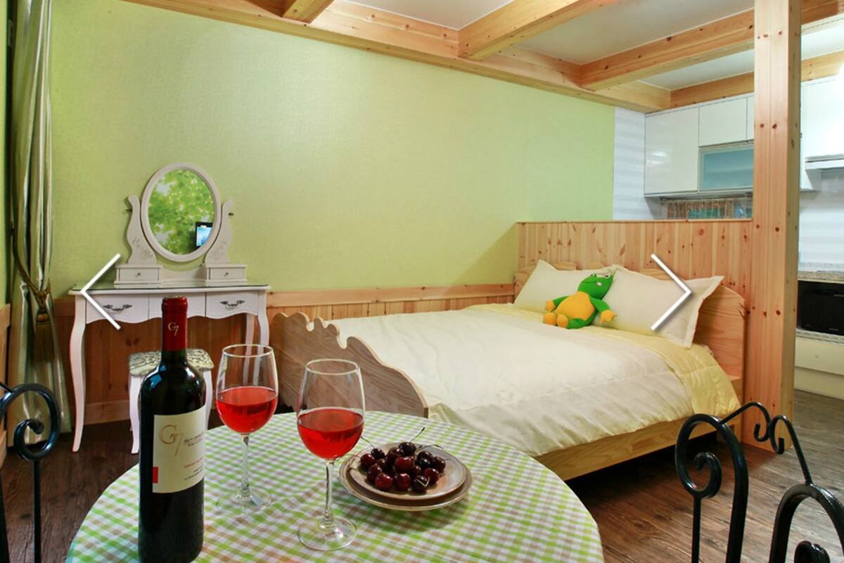 A bedroom with a bed, a small table with a bottle of wine and glasses, a mirror, and a green wall.