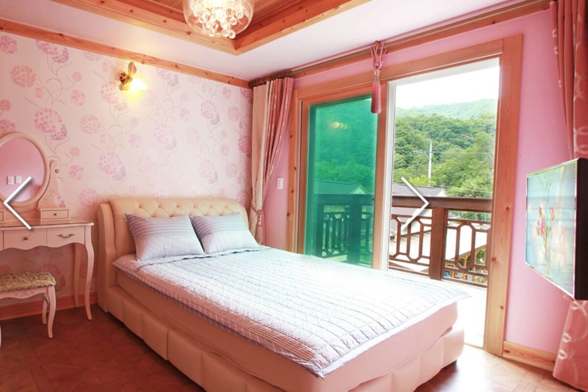 A bedroom with a bed, a dresser, a mirror, and a balcony with a view of greenery.