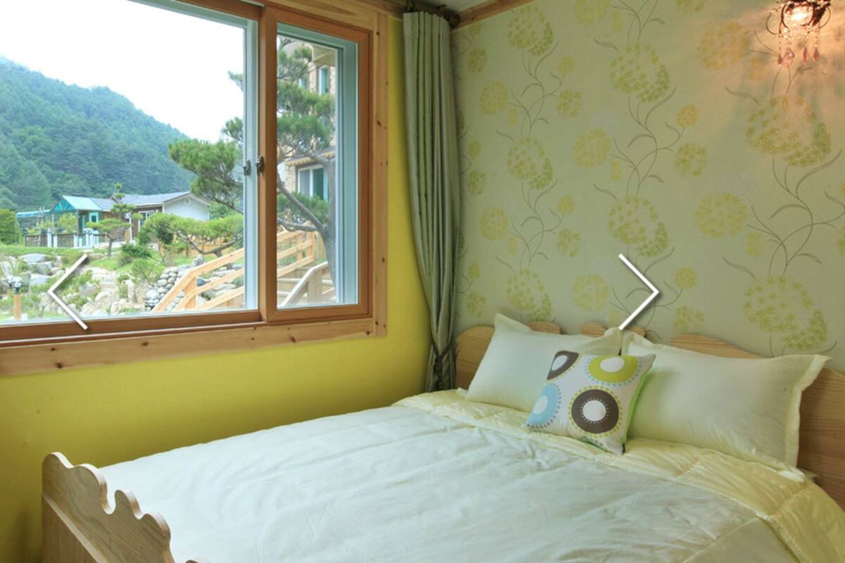 A bedroom with a bed, a window with a view of a mountain and a house, and a curtain.