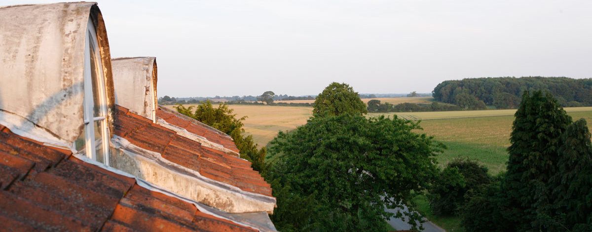 A view of a red-tiled roof with a window, surrounded by green trees and fields.