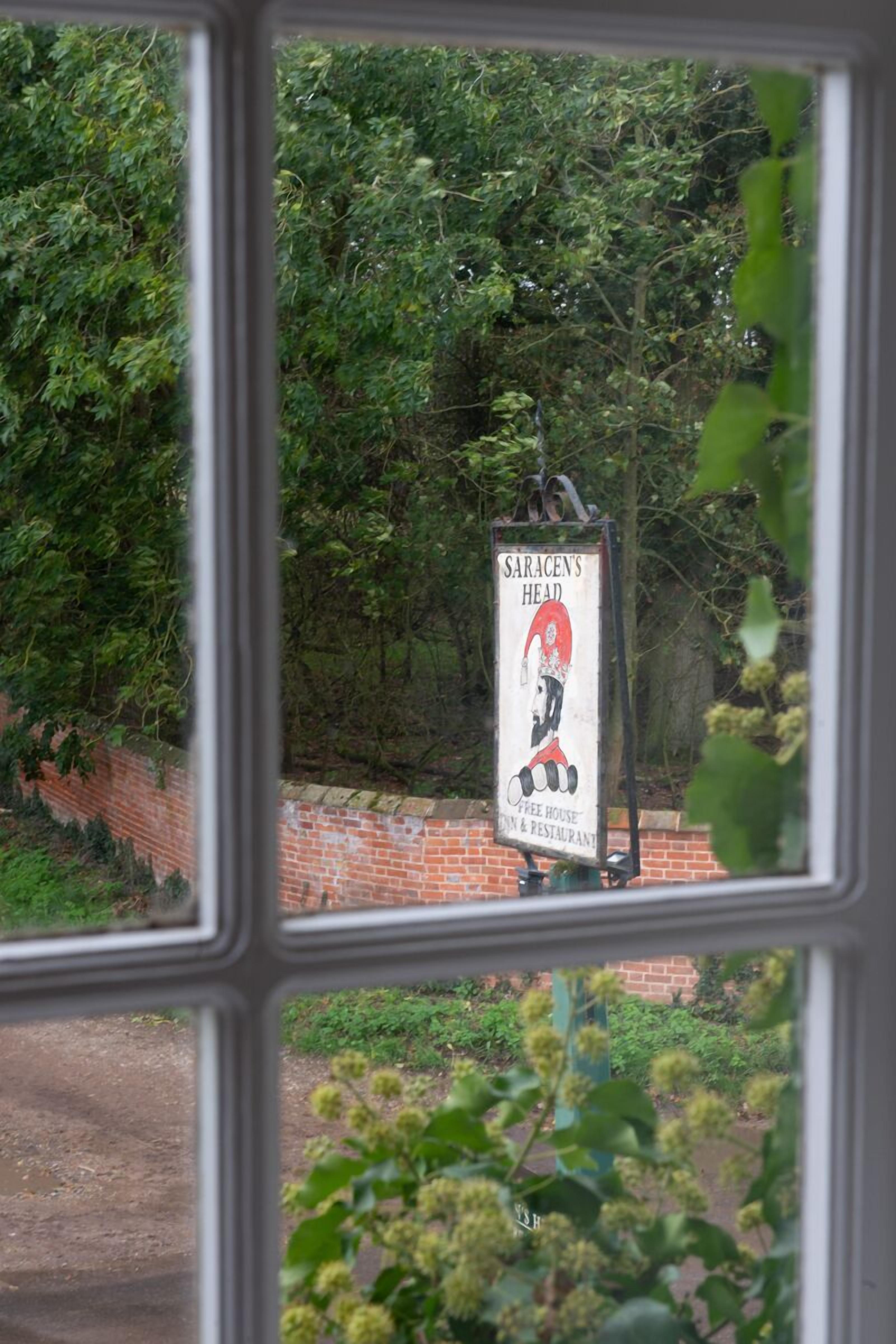 A sign for 'Sarasens Head' coffee house and restaurant with a red and white design, visible through a window with a brick wall and greenery in the background.