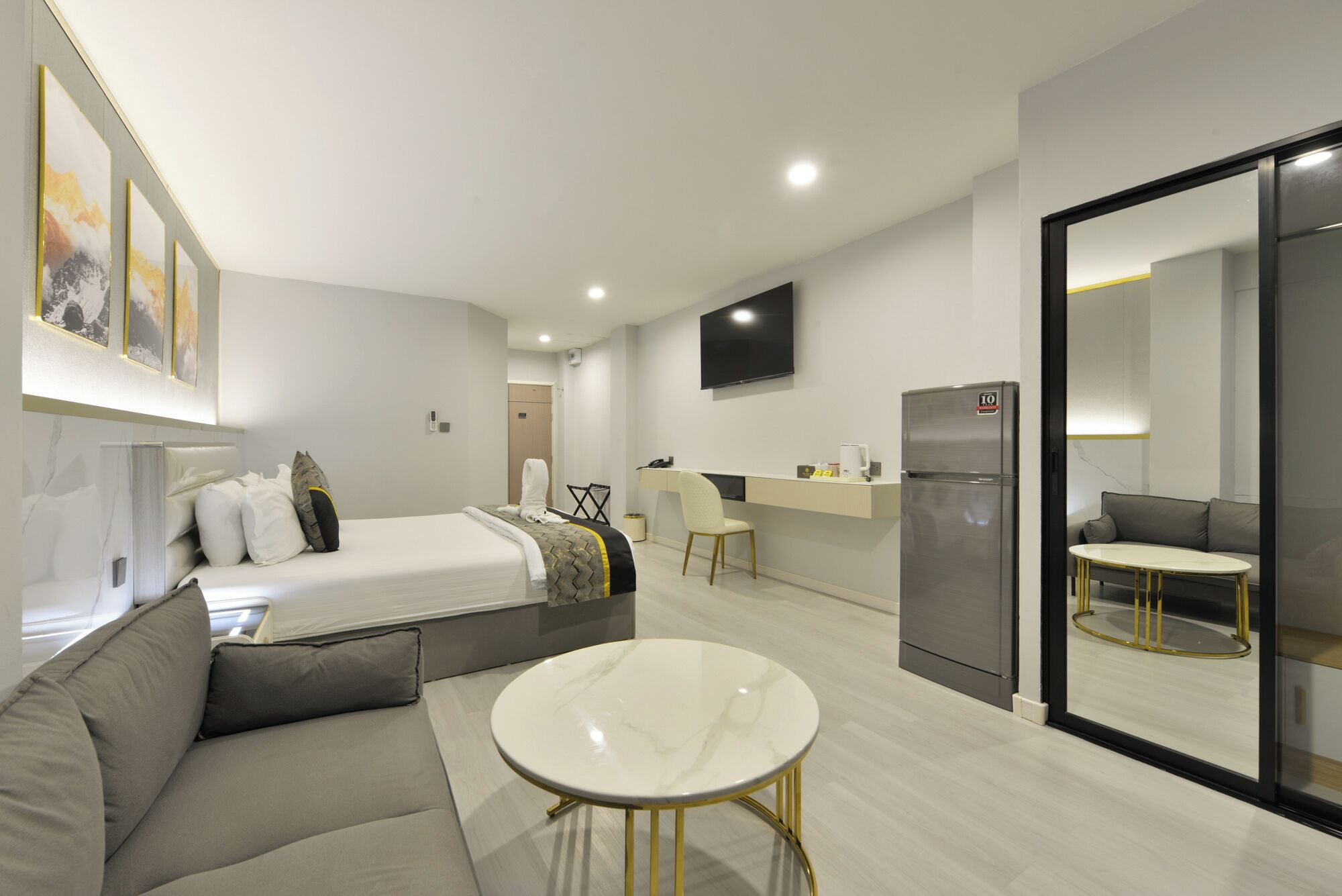 A modern hotel room with a bed, a sofa, a small round table, a kitchenette, and a TV mounted on the wall.