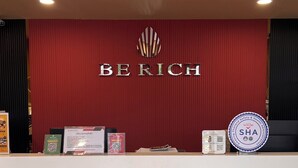 A reception desk with a red backdrop, the word "BERICH" prominently displayed, and a logo above it. There are informational signs, a QR code, and a circular sign indicating safety and health administration. - Berich Hotel (Tak)