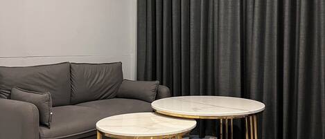 A modern living room with a grey sofa, two round tables with gold legs, and a large window with dark curtains.