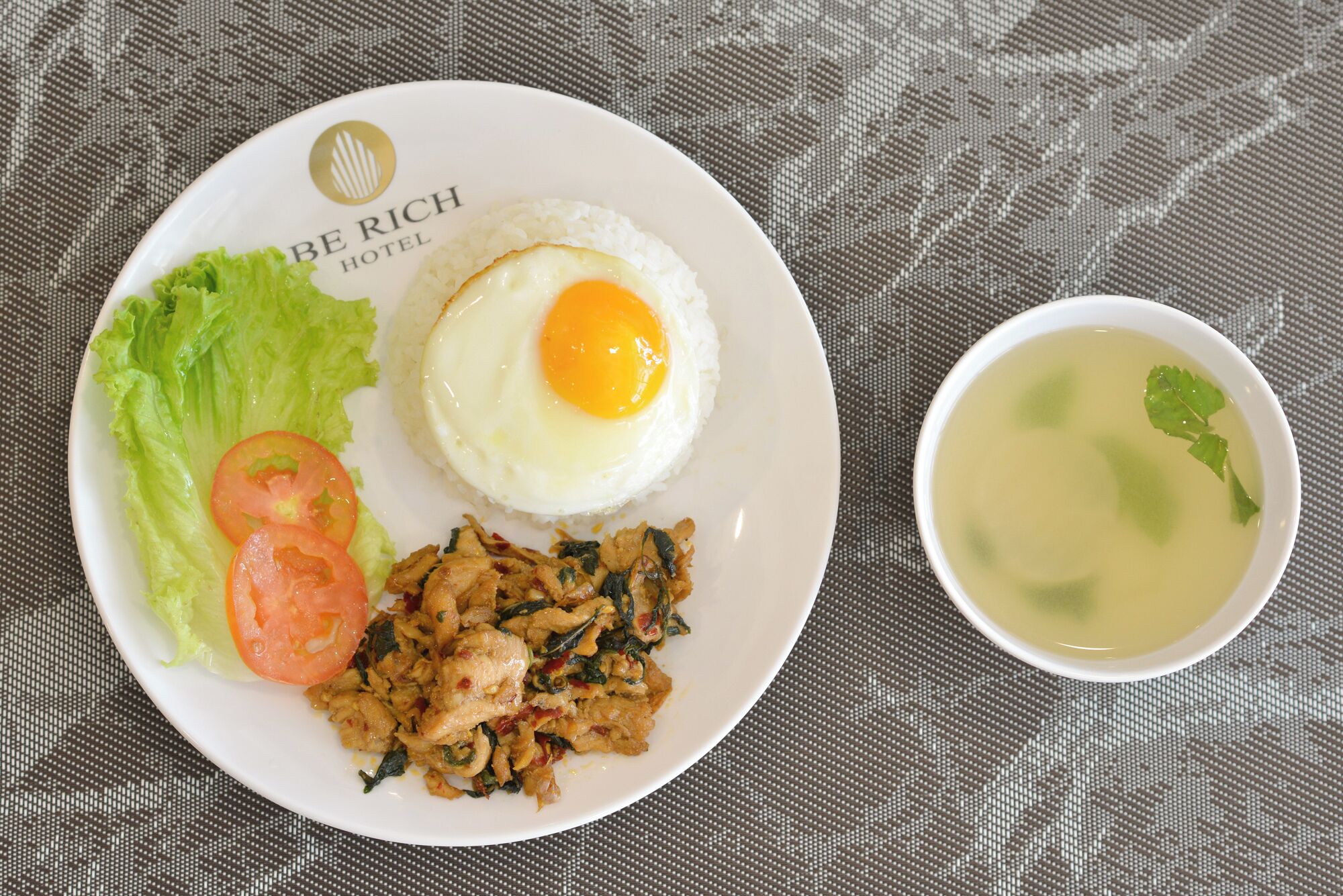 A plate with a fried egg, lettuce, tomato, and stir-fried chicken, and a bowl of soup.
