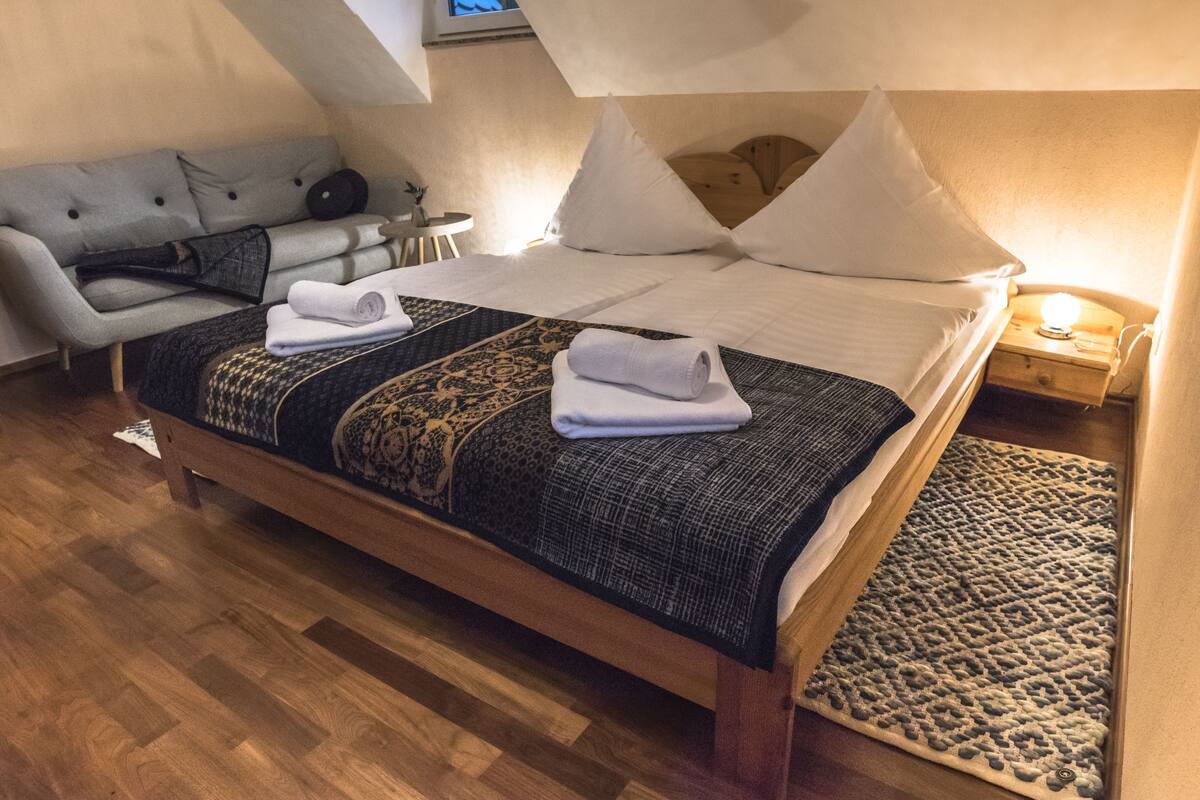 Ein ordentlich bezogenes Bett mit einem gemusterten Überwurf, ein graues Sofa und ein kleiner Tisch mit einer Lampe in einem Zimmer mit Holzboden.