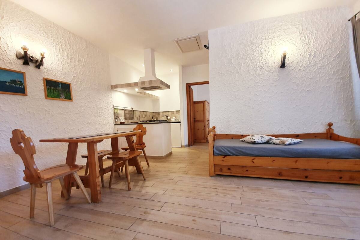 Um quarto acolhedor com uma mesa de jantar de madeira, cadeiras, uma cama e uma cozinha compacta ao fundo.