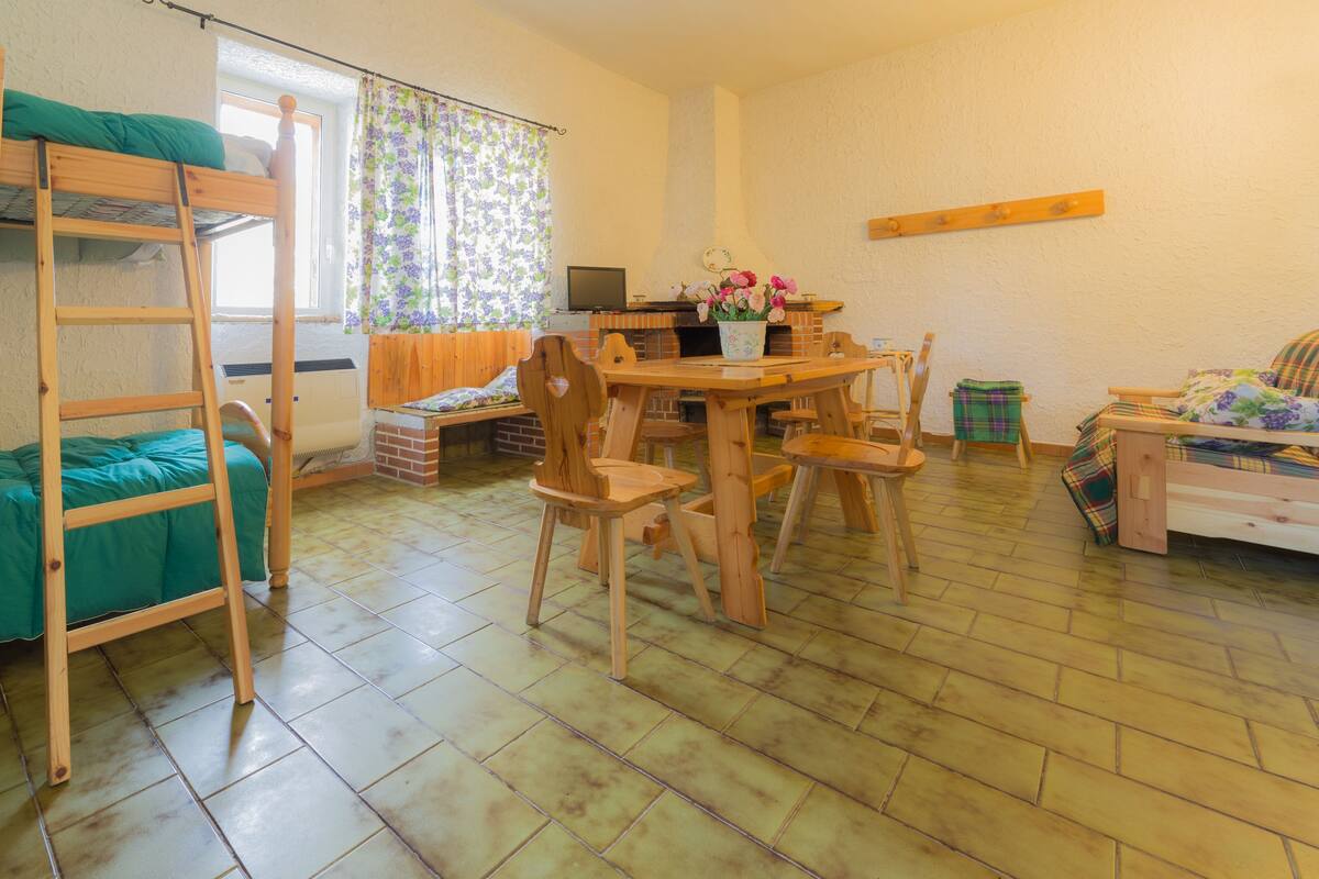 Um quarto aconchegante com uma cama de solteiro superior e inferior, uma mesa de jantar com cadeiras, uma mesinha com um vaso de flores, um sofá e uma estante de madeira.