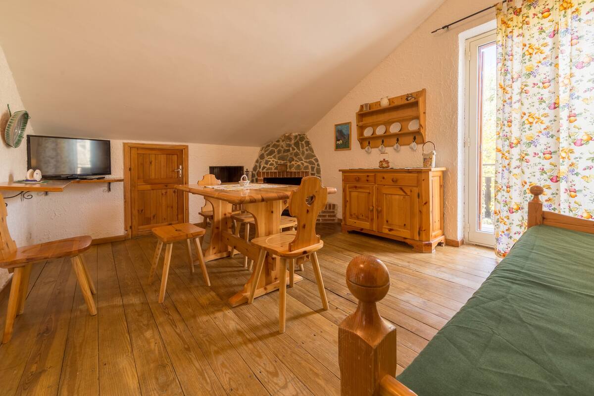 Um quarto aconchegante com uma mesa de jantar de madeira, cadeiras, uma lareira de pedra, armários de madeira, uma televisão e uma janela com cortinas florais.