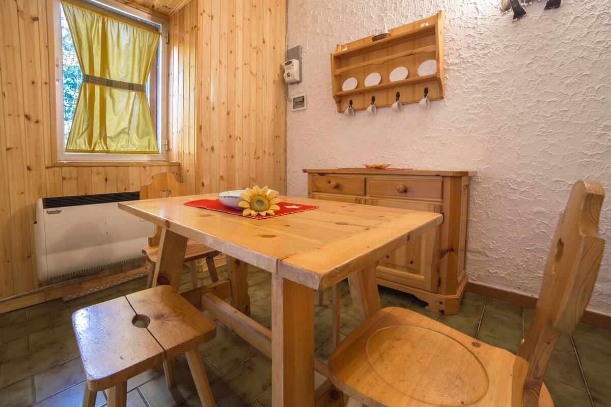 Interior de madeira com uma mesa, cadeiras, um radiador e uma estante com louças.