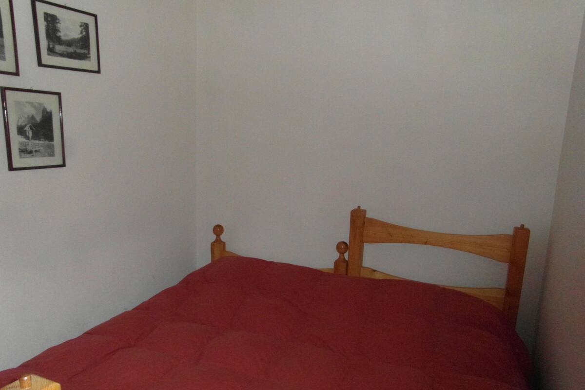 Quarto pequeno com uma cama de solteiro, cabeceira de madeira, colcha vermelha e duas fotografias emolduradas na parede.