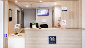 A hotel reception desk with a television displaying a cityscape, a potted plant, and a sign indicating no smoking. - ACE Hôtel Toulouse (Cornebarrieu)