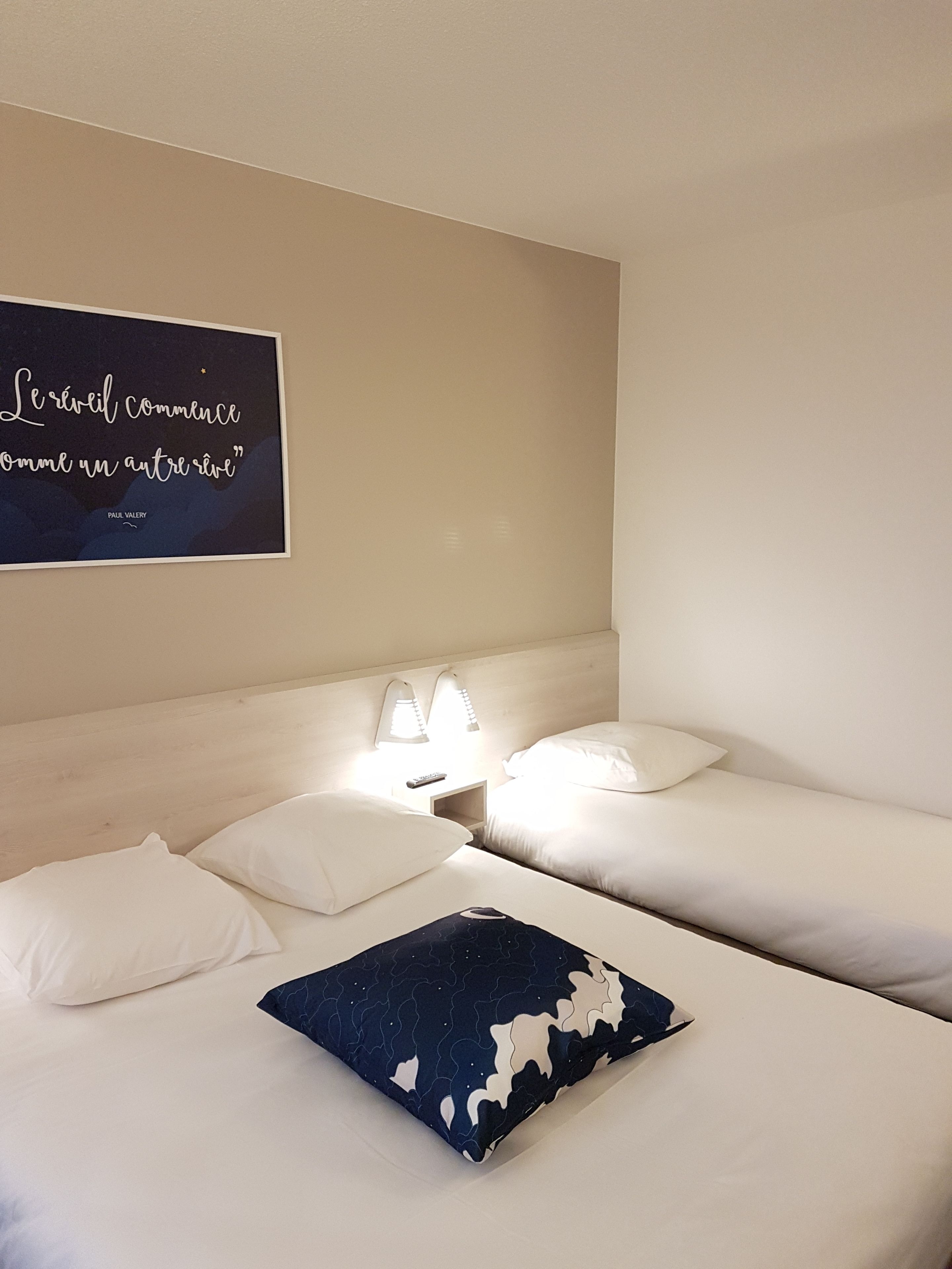 Una habitación de hotel con dos camas, una mesita de noche con lámpara y una cita en la pared.