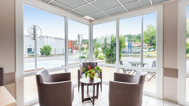A modern room with large windows, two grey armchairs, a small round table with a vase of flowers, and a view of the street and buildings outside. - ACE Hôtel Toulouse (Cornebarrieu)