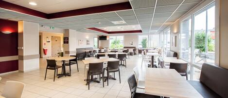 Una amplia cafetería con varias mesas y sillas, ventanas grandes y un techo alto con diseños de cuadrícula.
