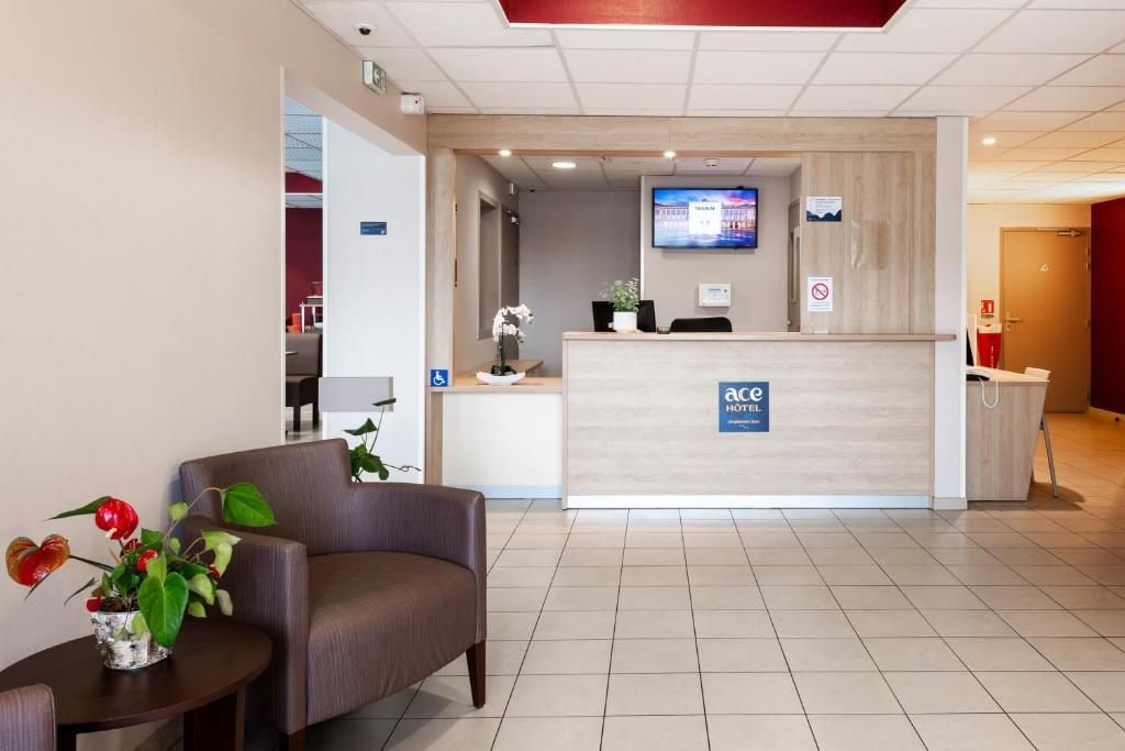 Zona de recepción de un hotel con mostrador, televisor de pantalla plana, área de estar con una planta y un cartel con el nombre del hotel.