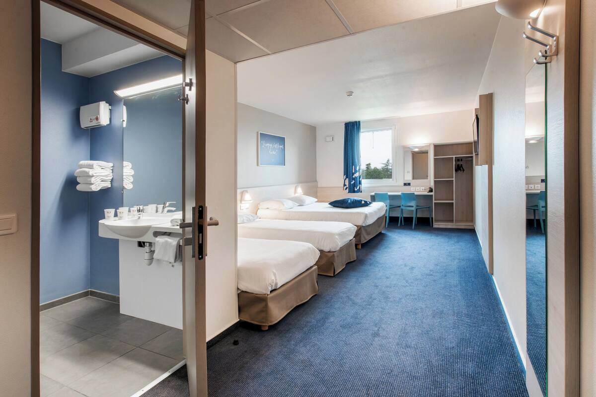 Une chambre d’hôtel avec trois lits, une salle de bain comprenant un lavabo et un porte-serviettes, ainsi qu’un bureau avec une chaise.
