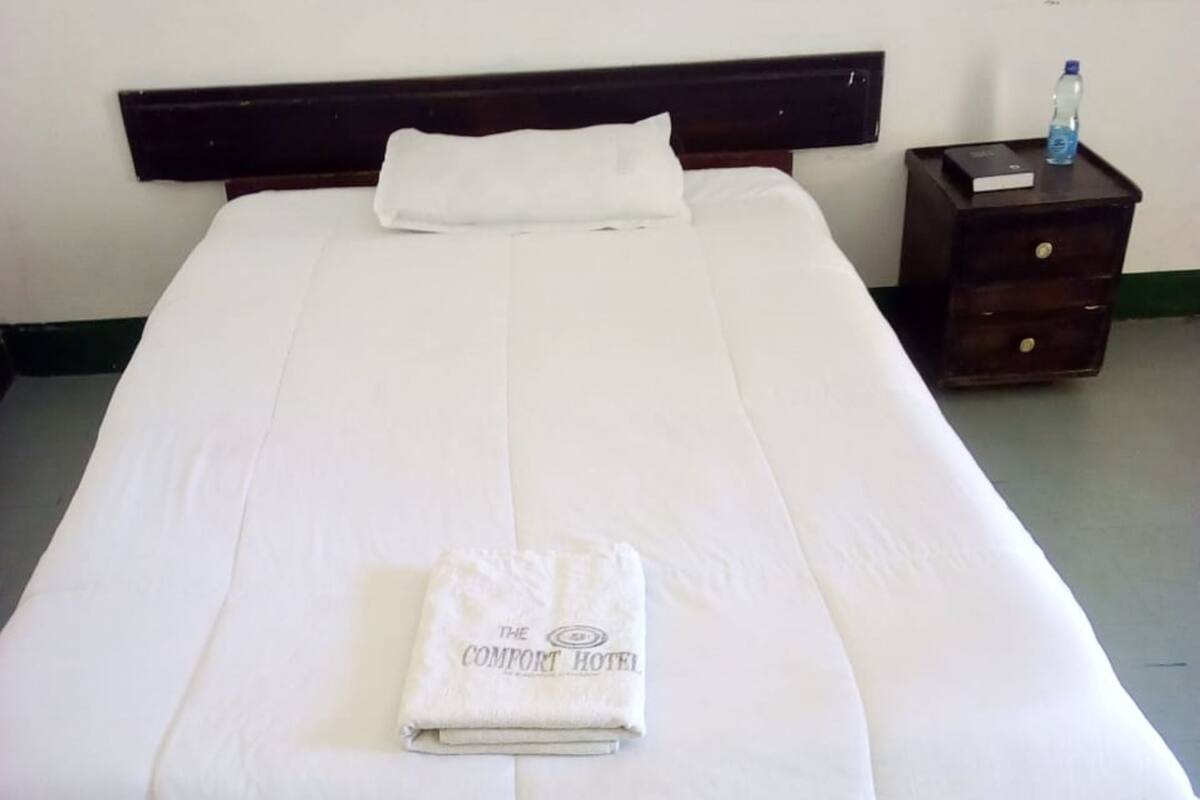 A neatly made bed with a folded towel, a pillow, and a nightstand with a bottle and a book.