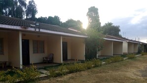 A row of single-story buildings with a tiled roof, a covered outdoor seating area, and a grassy lawn with sparse vegetation. - Family Comfort Hotel (Nanyuki)
