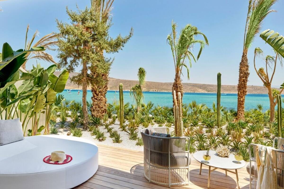 Una terraza de madera con una zona de estar circular, una pequeña mesa y sillas, con vistas a una playa con palmeras y un mar azul cristalino.