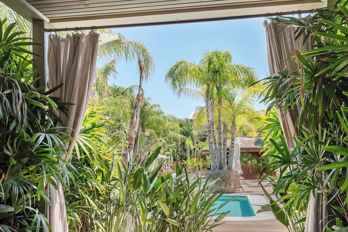 Un espacio de estar al aire libre techado con vista a la piscina, suelo de madera y plantas tropicales.