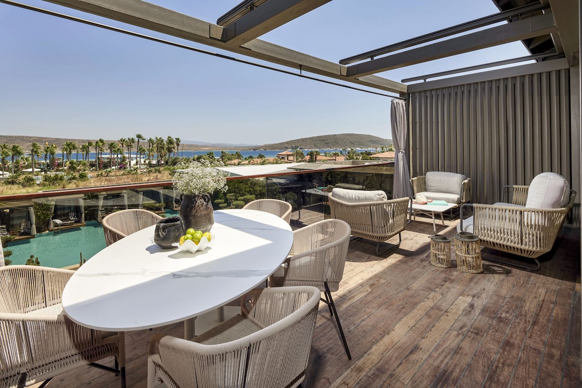 Un balcón con mobiliario de mimbre, una mesa blanca y vistas a una piscina y una playa.
