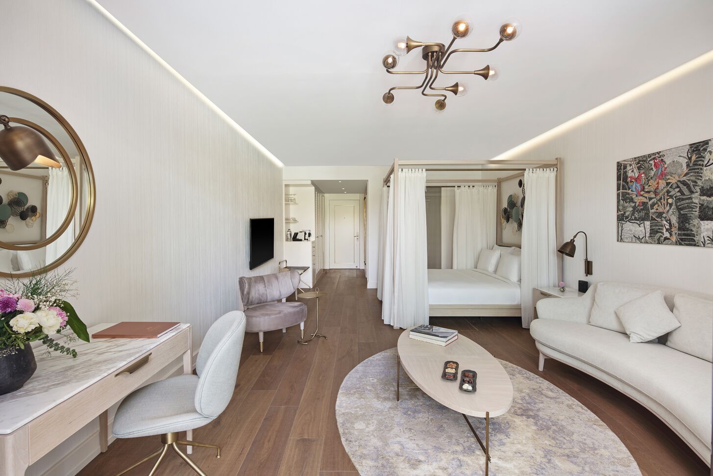 A modern bedroom with a four-poster bed, a sitting area with a sofa and armchair, a desk with a chair, and a large mirror.