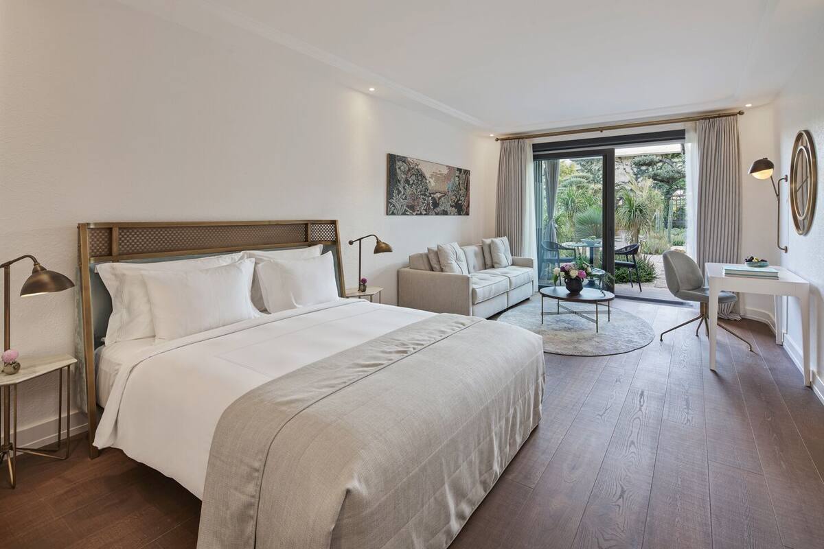 Un dormitorio amplio con una cama grande, una zona de estar con sofá y sillones, un escritorio y vistas al exterior a través de amplias ventanas.