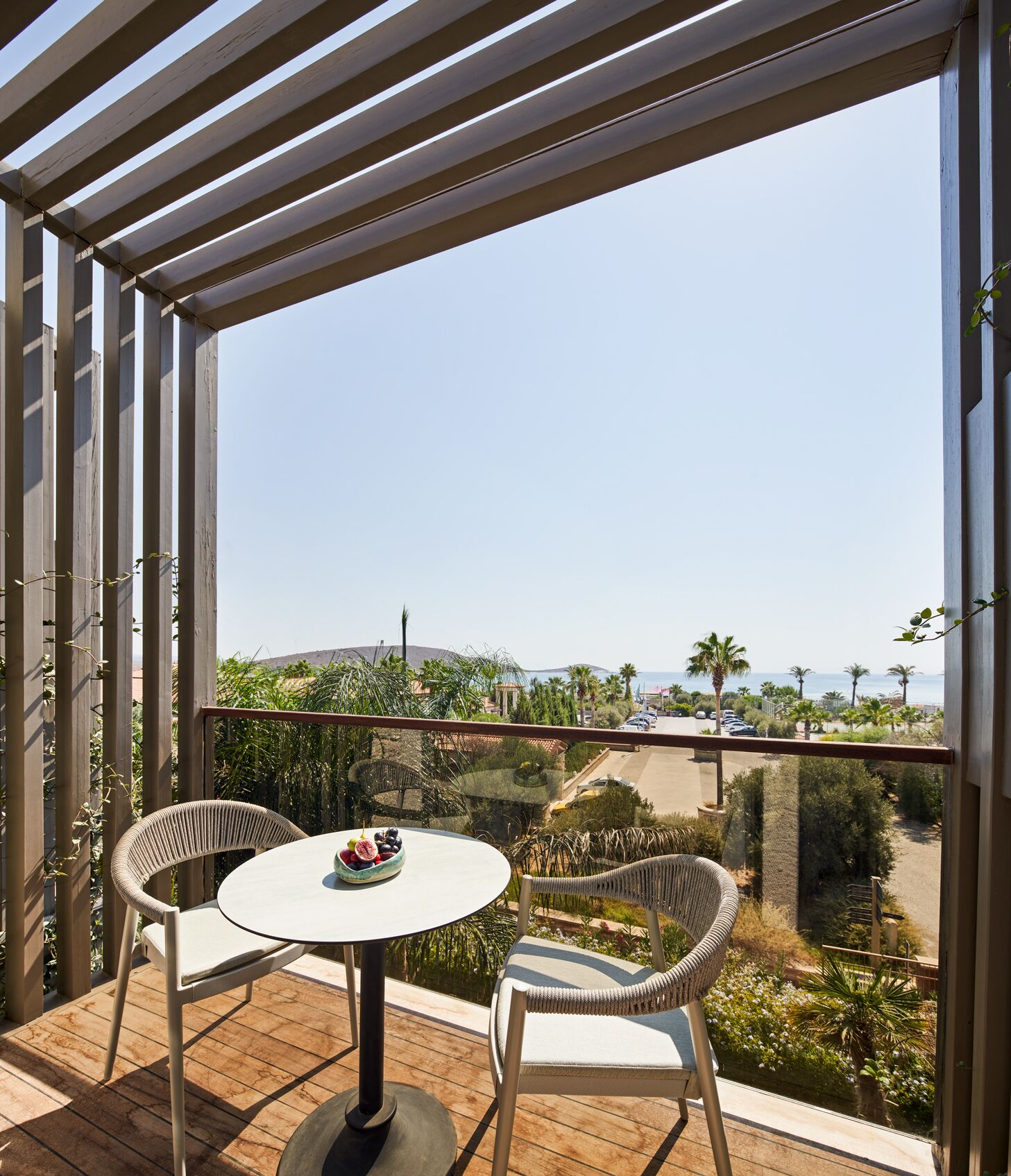 Un balcon offrant une vue sur un paysage côtier, avec deux fauteuils en osier et une petite table ronde sur laquelle sont disposés des fruits.