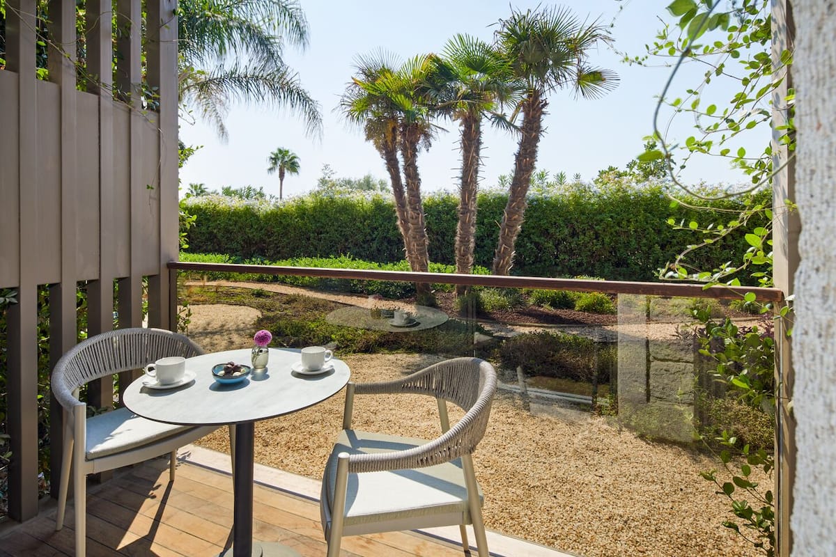 Un balcón con dos sillas de mimbre, una mesa redonda y barandilla de vidrio, con vista a un jardín con palmeras y seto.