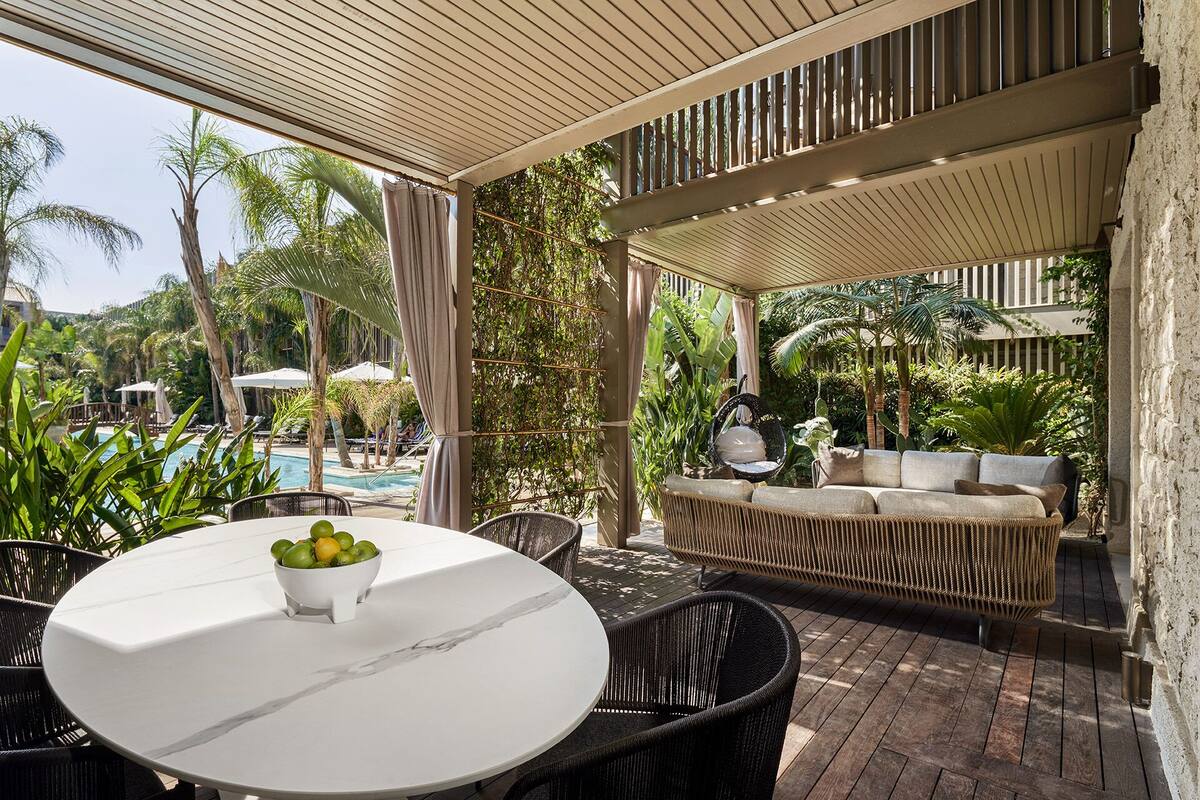 Un área de estar al aire libre techada con una mesa redonda y sillas, un sofá de mimbre y vista a una piscina y palmeras.